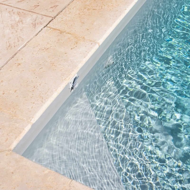 Close-up of pool edge with beige stone tiles and water reflection.
